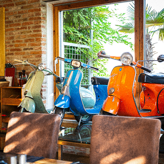 Drei Vintage-Roller in verschiedenen Farben sind in einem Raum mit einer Ziegelwand und einem großen Fenster ausgestellt, das Bäume und eine Palme draußen zeigt.