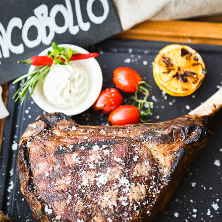 Ein Steak liegt auf einer schwarzen Oberfläche, garniert mit Kräutern, einer Chili, Tomaten, einer halben Zitrone und einer Schüssel Dip. Ein Schild mit dem Wort 'Ancobollo' ist im Hintergrund.