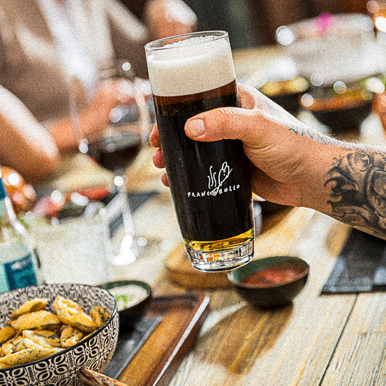 Eine Person, die ein Glas dunkles Bier hält, mit einer Tätowierung am Arm, sitzt an einem Tisch mit anderen.