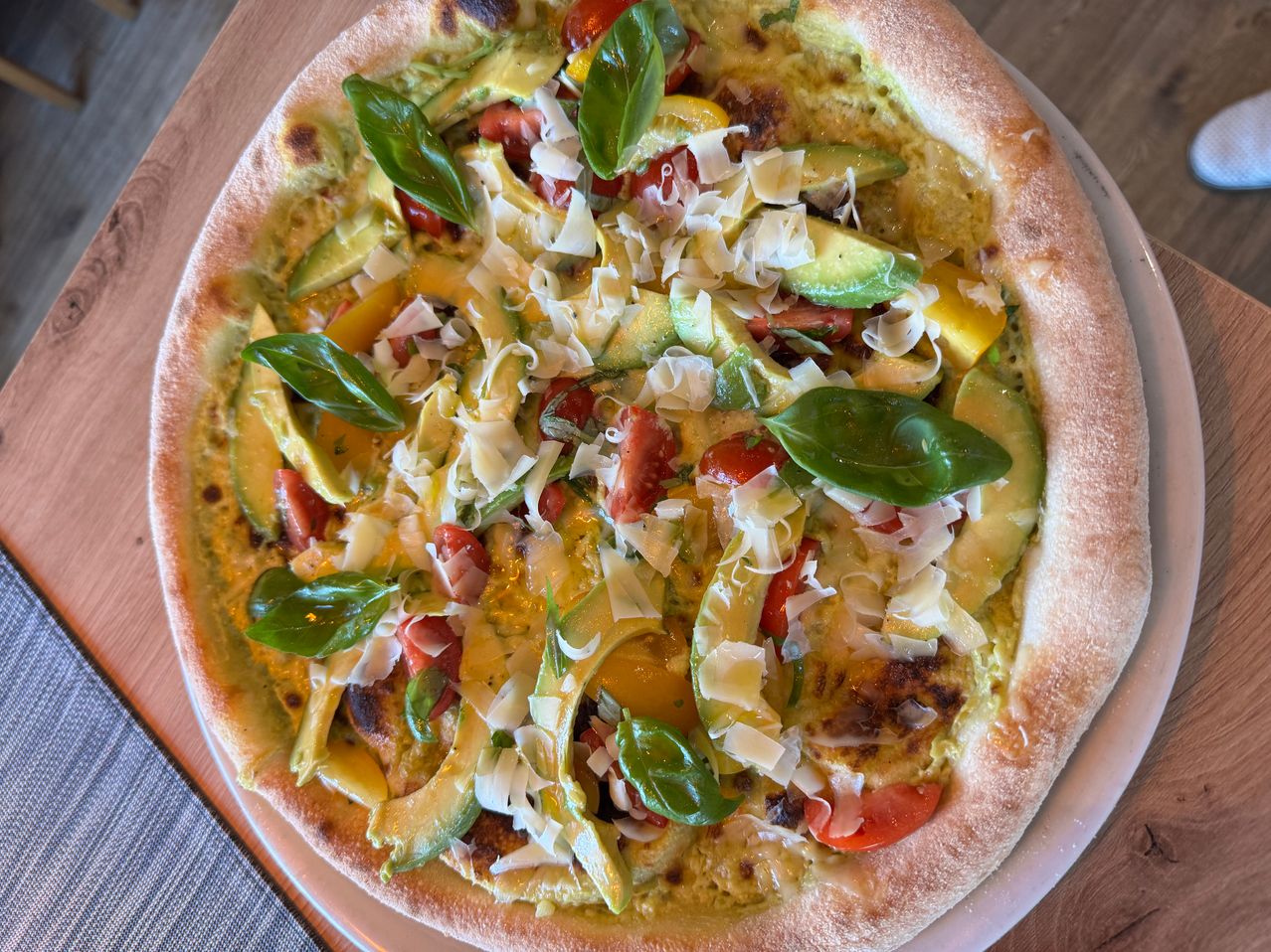 Eine Pizza mit Avocado, Tomaten, Basilikumblättern und geriebenem Käse. Sie hat einen goldenen Rand und steht auf einem weißen Teller.