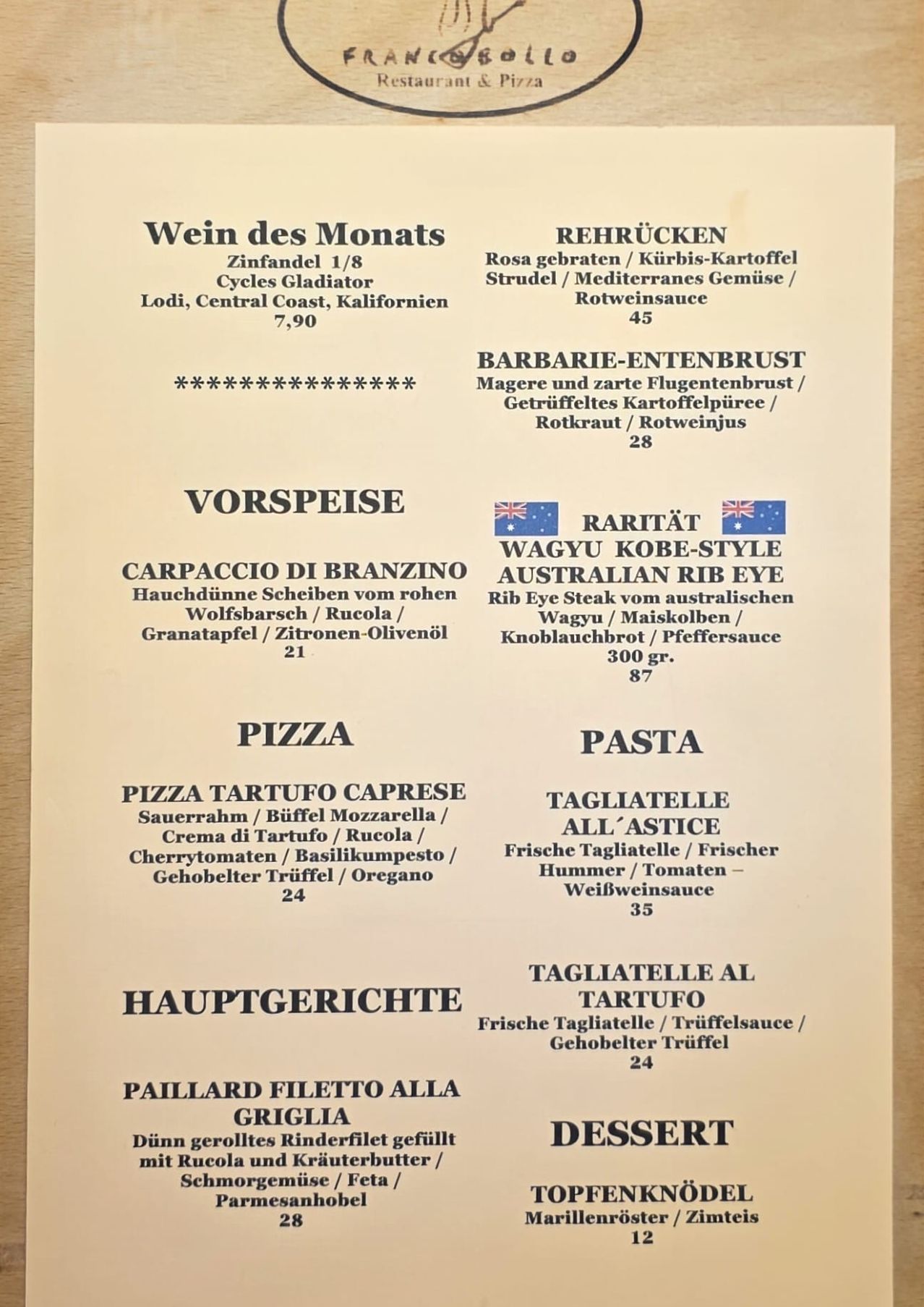 Eine Speisekarte zeigt einen Wein des Monats, roten Zinfandel. Vorspeisen umfassen Carpaccio di Branzino und Wagyu Kobe-Style Australian Rib Eye. Pasta-Optionen sind Tagliatelle All'Astice und Tagliatelle al Tartufo.