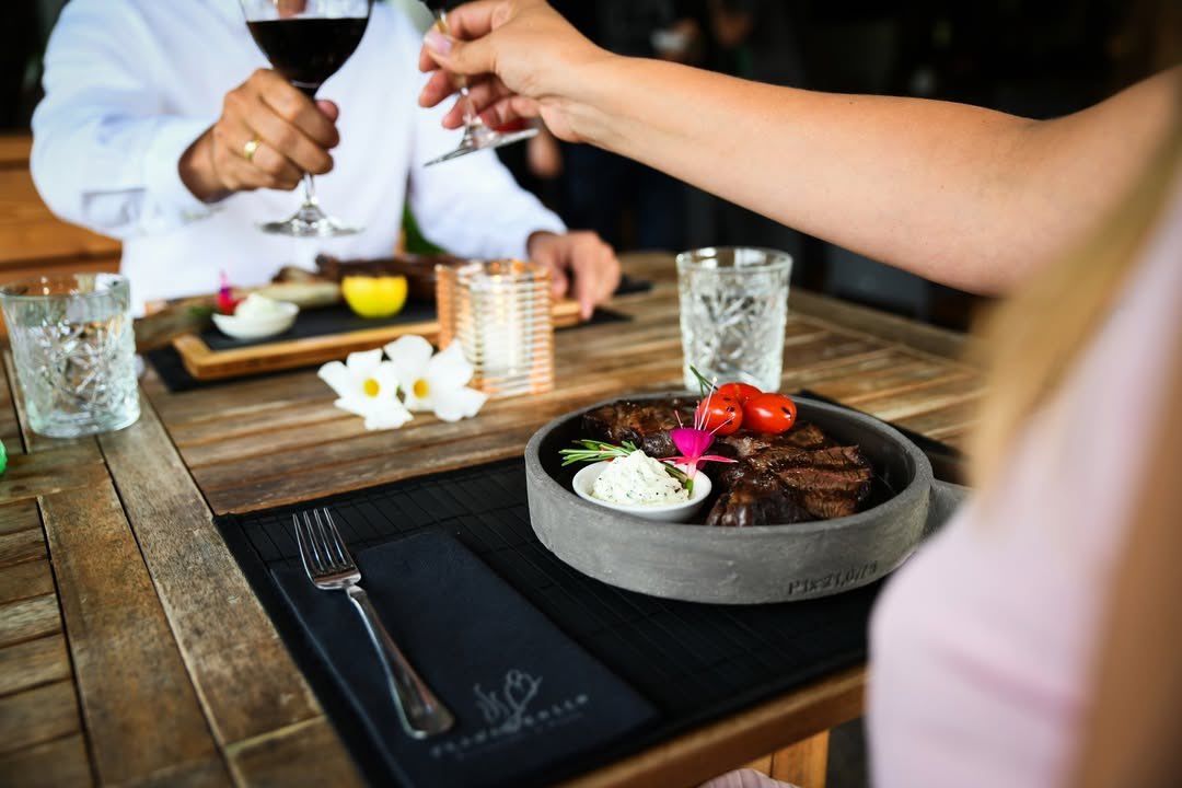 Zwei Personen, die mit Weingläsern anstoßen, an einem Tisch mit einem Steakteller und Garnierungen, zusammen mit einer Gabel auf einem schwarzen Tuch.