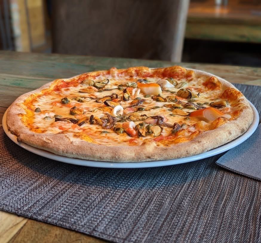 Eine runde Pizza mit Meeresfrüchten, Käse und roter Sauce auf einem weißen Teller steht auf einem Holztisch.