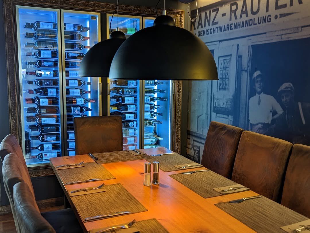 Ein Essbereich mit einem Holztisch, der mit Platzkarten und Besteck gedeckt ist. Dahinter steht ein Weinregal mit blau leuchtenden Lichtern. An der Wand steht ein großes Foto eines Mannes.