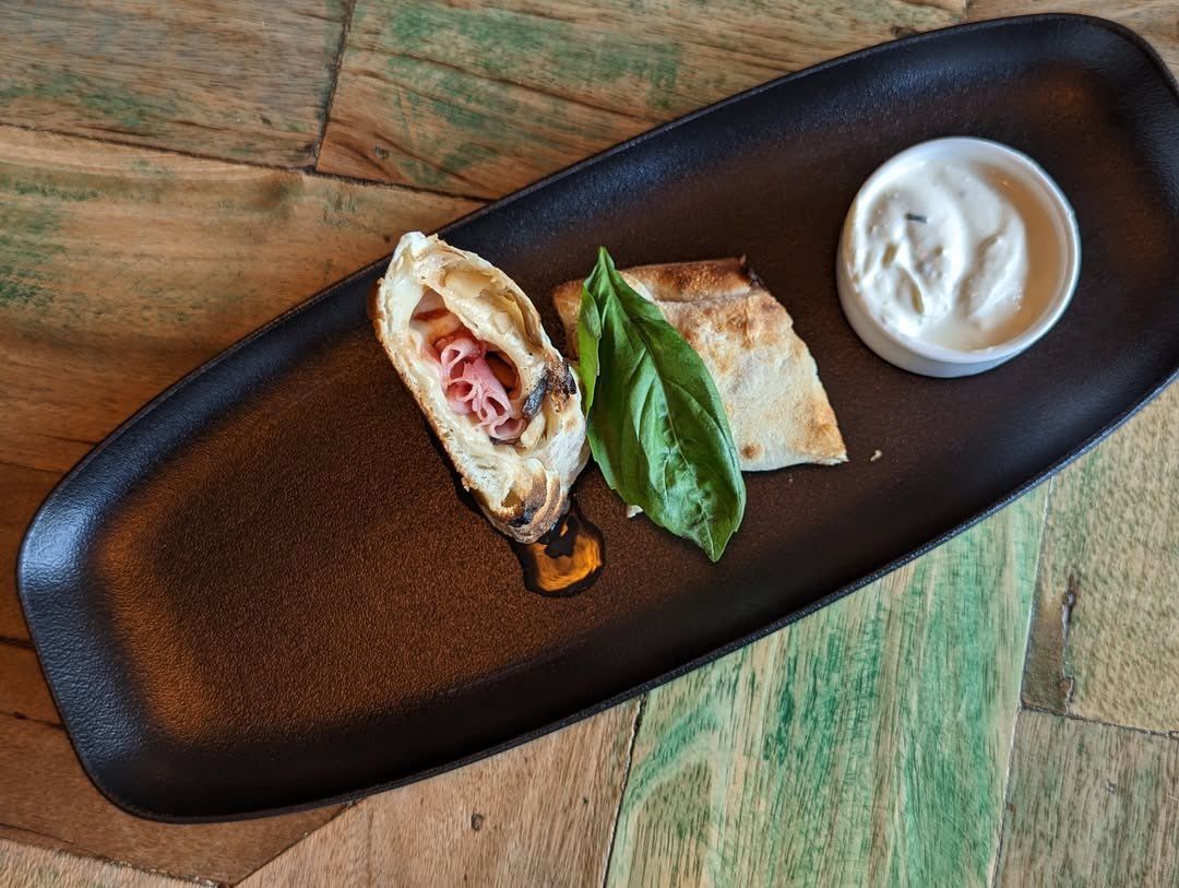 Ein Teller mit einem getoasteten Sandwich, einem Basilikumblatt und einer kleinen Schüssel mit weißer Sauce auf einem Holztisch.