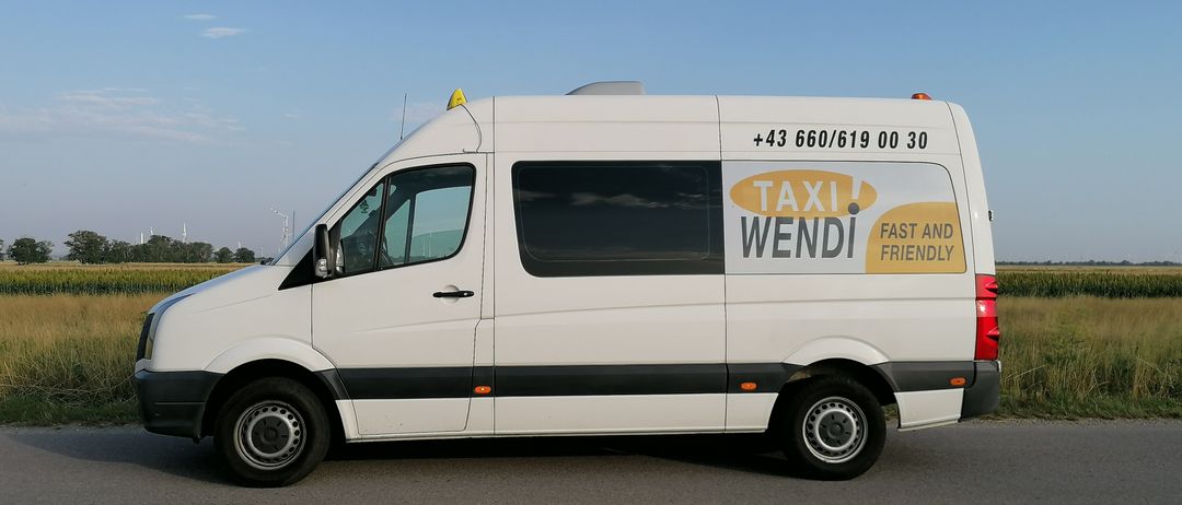 Ein weißer Lieferwagen mit einem gelben Schild, auf dem 'Taxi! WENDI. Schnell und freundlich' steht, ist auf einer Straße geparkt.