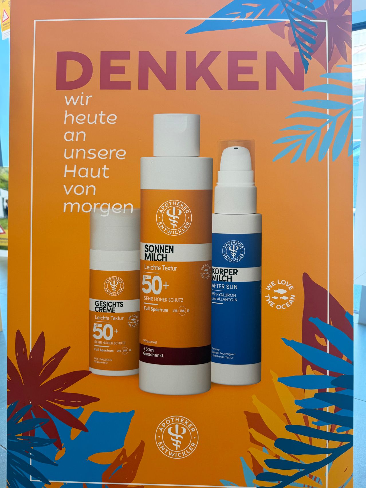 Ein orangefarbener Plakat zeigt drei Sonnenschutzflaschen, zwei mit 'Gesichts Creme 50+' und eine mit 'Körper Milch After Sun.' Die Flaschen haben weiße Kappen und sind auf einem orangefarbenen Hintergrund angeordnet. Das Plakat hat ein blaues und weißes Blattdesign und Text, der 'Denken wir heute an unsere Haut von morgen' lautet.