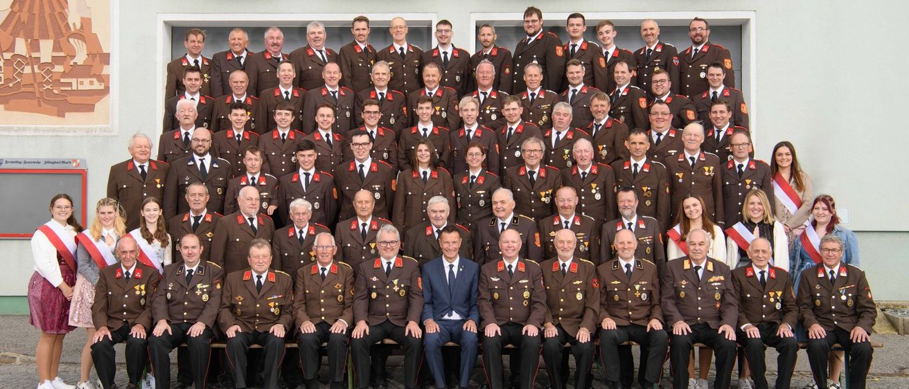 Eine große Gruppe von Menschen in Uniformen posiert für ein Foto vor einem Gebäude mit Blumen und feiert 150 Jahre FF Zillingdorf-Markt im Jahr 2024.