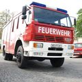 Ein roter Feuerwehrwagen mit der Aufschrift 'FEUERWEHR' steht auf einer Kopfsteinpflasterstraße. Er hat ein Kennzeichen mit der Aufschrift 'WB FFZM1'. Ein rotes Auto ist dahinter geparkt.