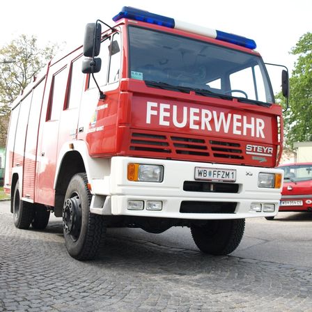 Ein roter Feuerwehrwagen mit der Aufschrift 'FEUERWEHR' steht auf einer Kopfsteinpflasterstraße. Er hat ein Kennzeichen mit der Aufschrift 'WB FFZM1'. Ein rotes Auto ist dahinter geparkt.