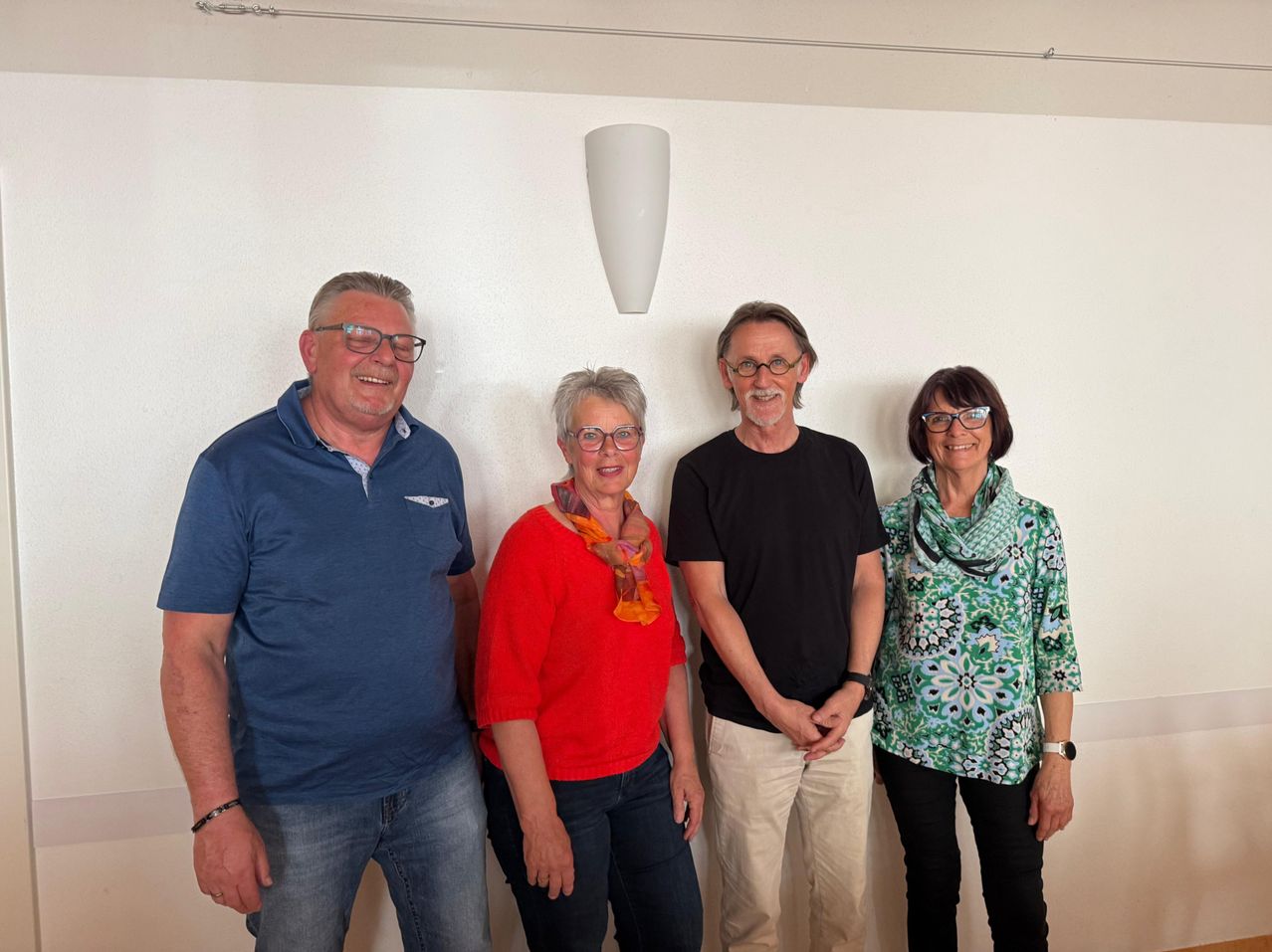 Fünf Erwachsene stehen lächelnd gegen eine weiße Wand. Ein Mann in Blau links, eine Frau in Rot in der Mitte, ein Mann in Schwarz neben ihr und zwei Frauen in Grün und Schwarz rechts.