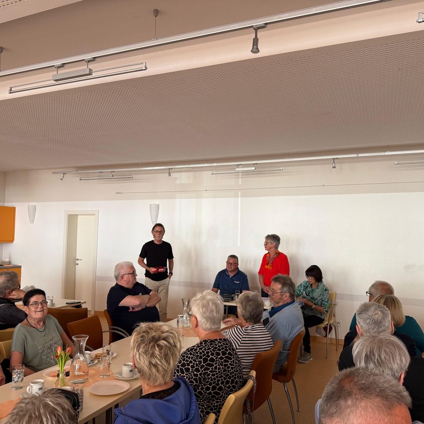 Eine Gruppe älterer Menschen sitzt um einen Tisch in einem Besprechungsraum herum. Ein Mann steht in der Nähe der Wand und eine Frau steht hinter dem Tisch. Es gibt Lampen an der Decke. Es gibt eine Tür auf der linken Seite des Raumes. Es gibt einen Schreibtisch vor der Tür. Es gibt Vasen auf dem Tisch.