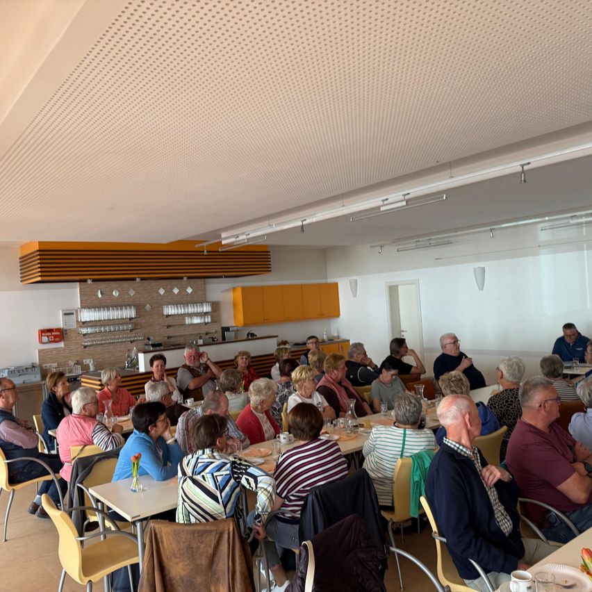 Ein Speisesaal voller älterer Menschen, die um Tische herum sitzen und sich unterhalten. Der Raum hat weiße Wände, Holzböden und orangefarbene Schränke. Ein Tisch mit einer Blumenvase steht in der Mitte.