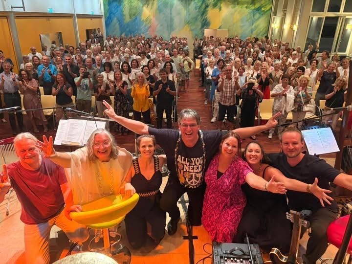 Eine Gruppe von Musikern und Zuschauern feiert gemeinsam in einem großen Raum mit Musikinstrumenten und einer farbenfrohen Wandmalerei im Hintergrund.