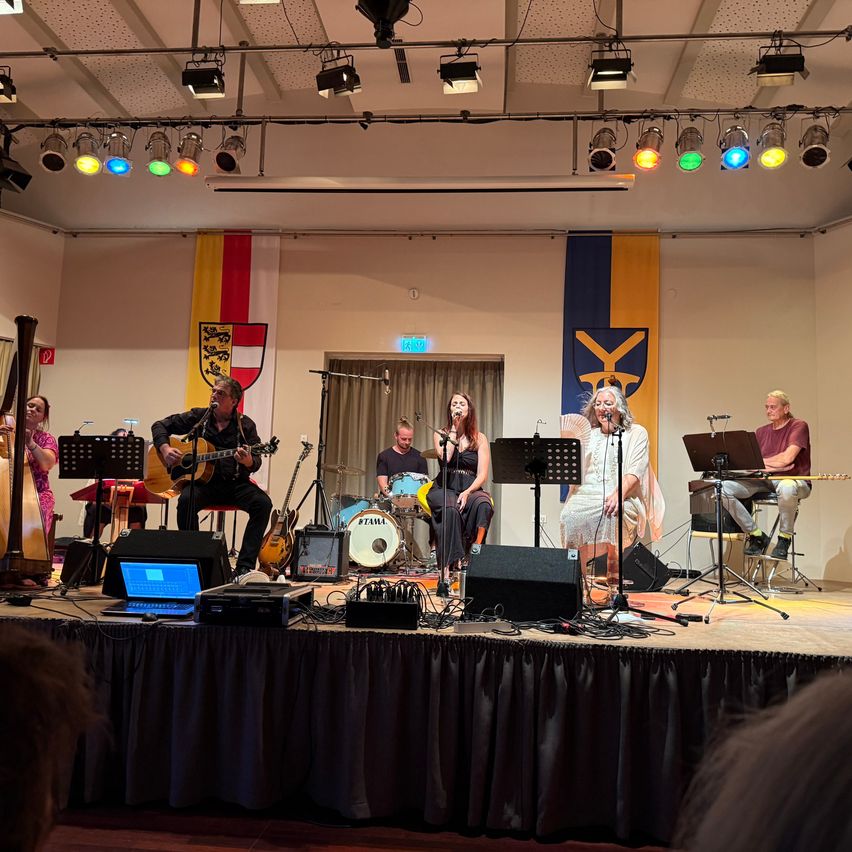 Eine Band spielt auf der Bühne mit verschiedenen Instrumenten, darunter Gitarren, Schlagzeug und Harfe, unter bunten Bühnenlichtern.