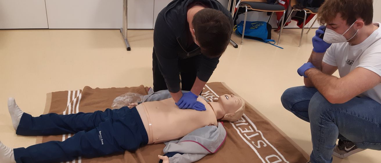 Eine Person mit blauen Handschuhen führt eine Herz-Lungen-Wiederbelebung an einem Mannequin durch, das eine Jacke trägt und auf einem braunen Teppich liegt, während eine andere Person in der Nähe steht. Im Hintergrund befinden sich ein weißer Tisch und Stühle.