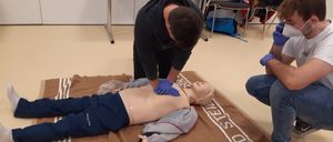 Eine Person mit blauen Handschuhen führt eine Herz-Lungen-Wiederbelebung an einem Mannequin durch, das eine Jacke trägt und auf einem braunen Teppich liegt, während eine andere Person in der Nähe steht. Im Hintergrund befinden sich ein weißer Tisch und Stühle.