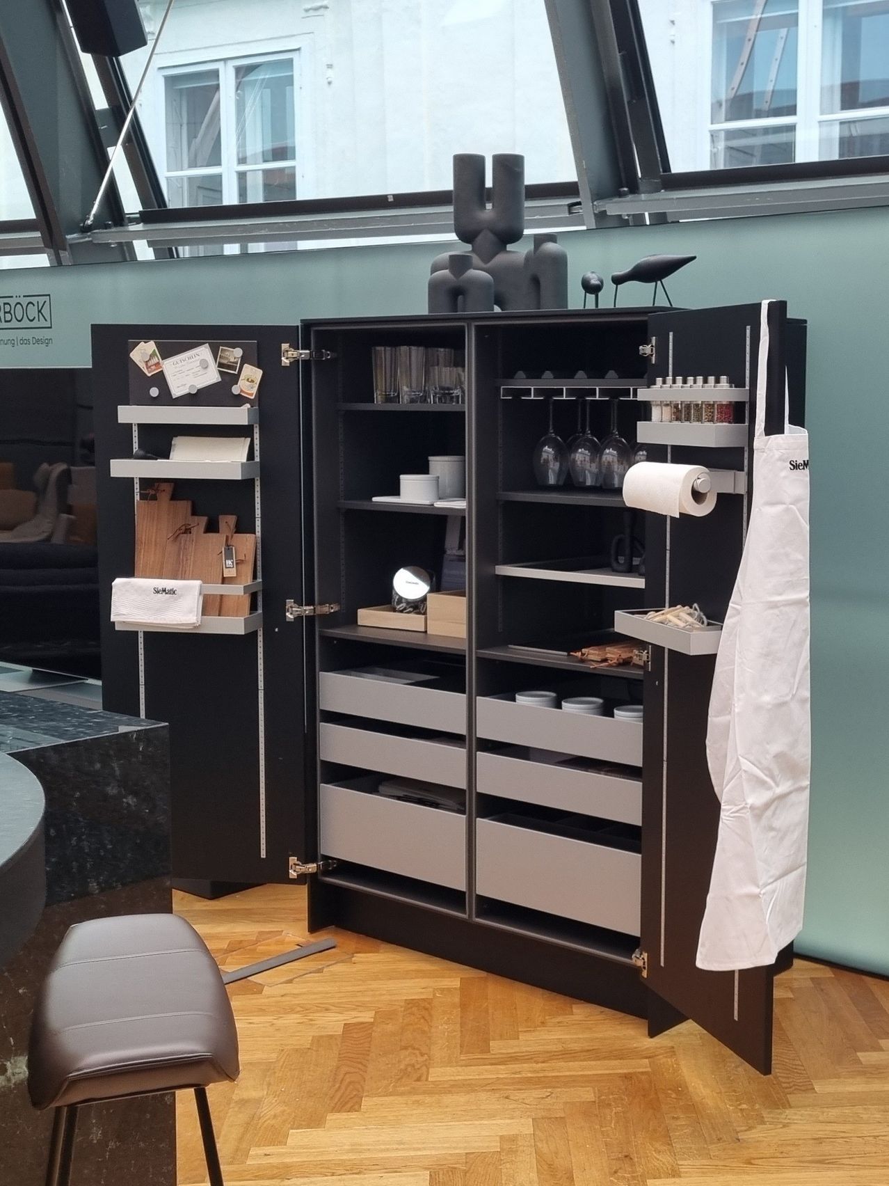 Ein moderner, schlanker schwarzer Küchenschrank mit mehreren Schubladen und Regalen, der verschiedene Küchenutensilien und Geschirr zeigt. Eine Schürze hängt auf der rechten Seite.