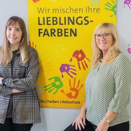 Zwei Frauen stehen neben einem farbenfrohen Schild mit Handabdrücken, lächeln und posieren für ein Foto. Das Schild steht auf Deutsch 'Wir mischen Ihre Lieblingsfarben'.