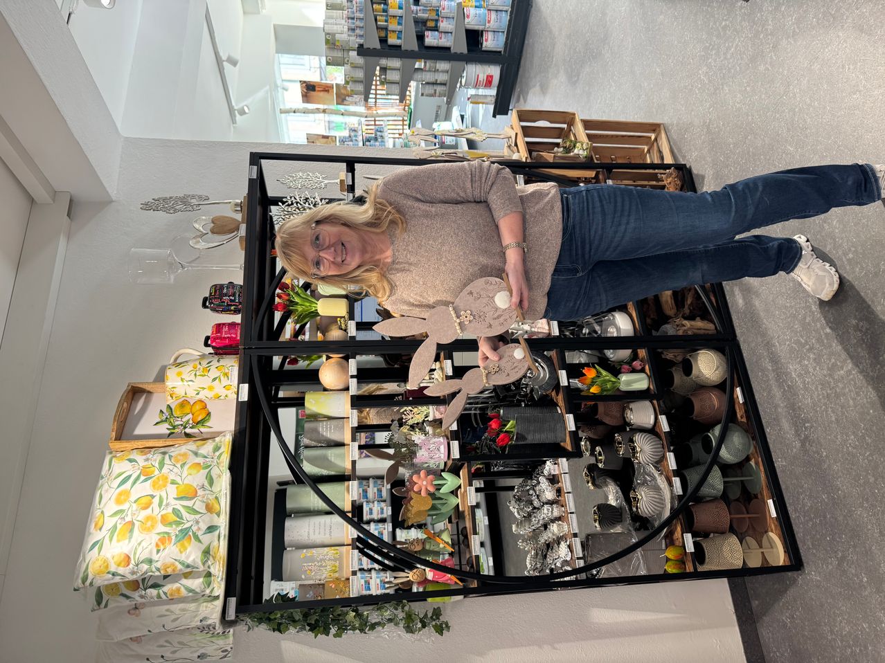 A woman stands in a shop, smiling, holding a decorative bunny. Shelves around her are filled with various items.