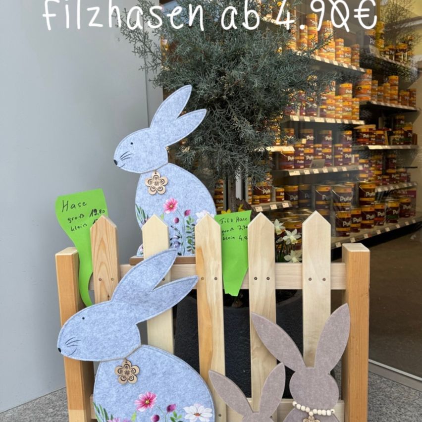 Three felt bunnies in different sizes, each with flowers and a sign, stand in front of a wooden fence. Behind the fence, a plant and a shop window with jars are visible. The text 'Filzhasen ab 4.99€' is written on the image.