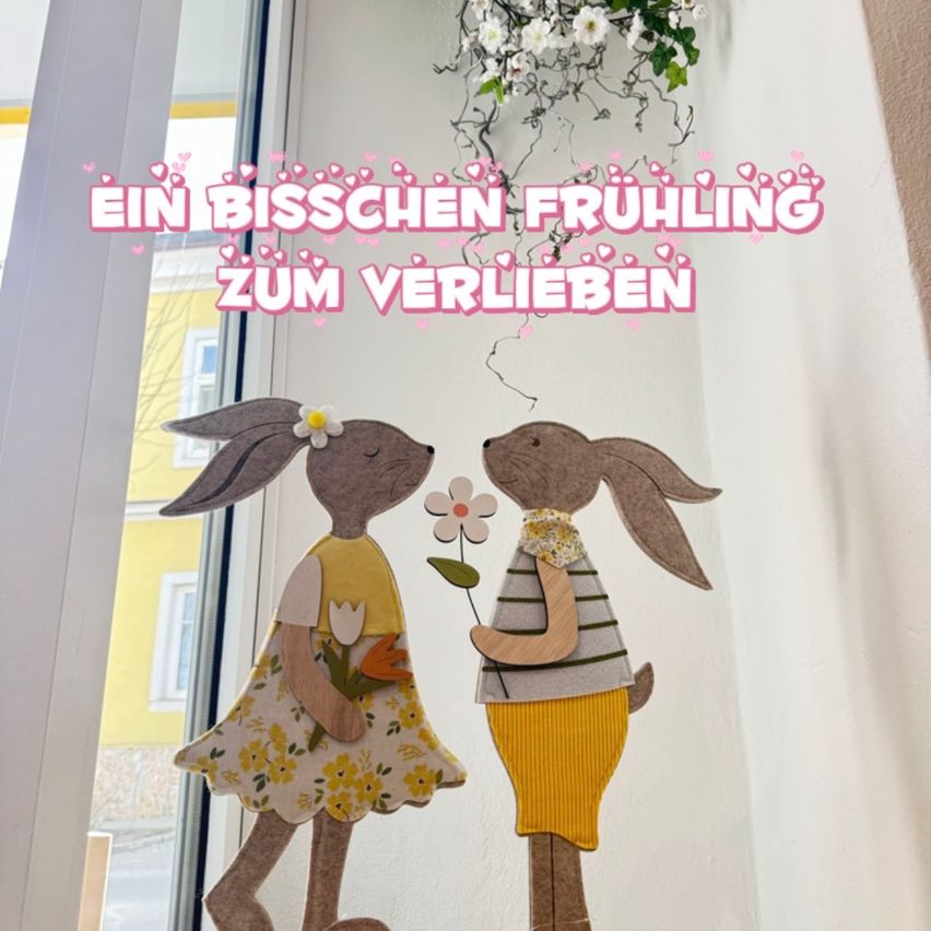 Two rabbit-shaped paper cutouts stand by a window with a floral decoration above. The cutouts hold flowers and the text reads 'EIN BISCHEN FRUHLING ZUM VERLIEBEN'.