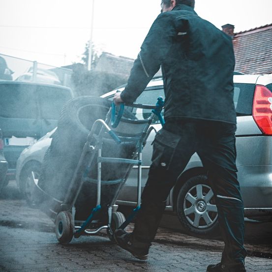 Ein Mann schiebt einen Wagen mit zwei Reifen durch einen Parkplatz, mit einem Haus im Hintergrund.