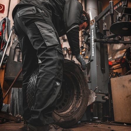 Ein Mann in einer Garage beugt sich über einen Reifen, trägt schwarze Hose und Handschuhe.