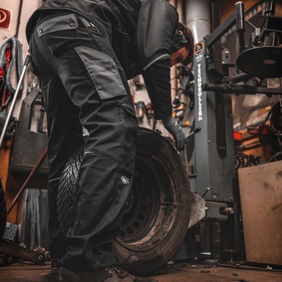 Ein Mann in einer Garage beugt sich über einen Reifen, trägt schwarze Hose und Handschuhe.
