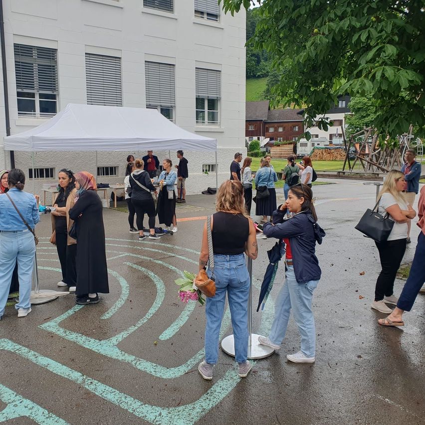 Eine Gruppe von Menschen steht draußen in der Nähe eines weißen Zelts, mit einem großen Gebäude dahinter. Einige schauen auf ein Spiraldesign auf dem Boden. Eine Frau hält einen Blumenstrauß und einen Regenschirm.