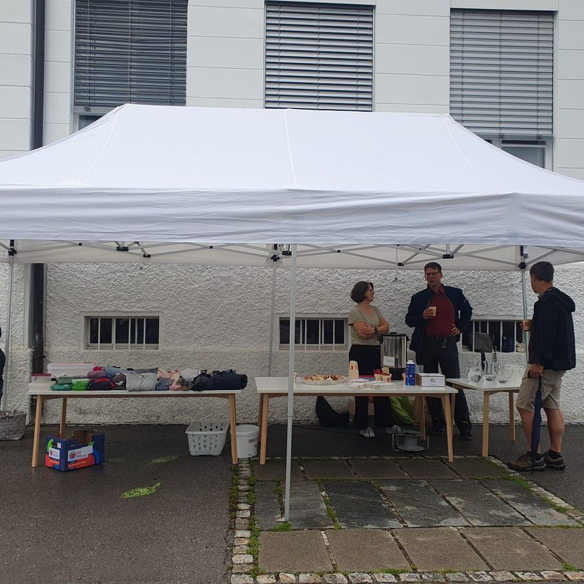 Drei Personen stehen unter einem Zelt in der Nähe eines weißen Gebäudes. Zwei Tische halten Essen, Kleidung und andere Gegenstände. Eine Person hält einen Regenschirm.