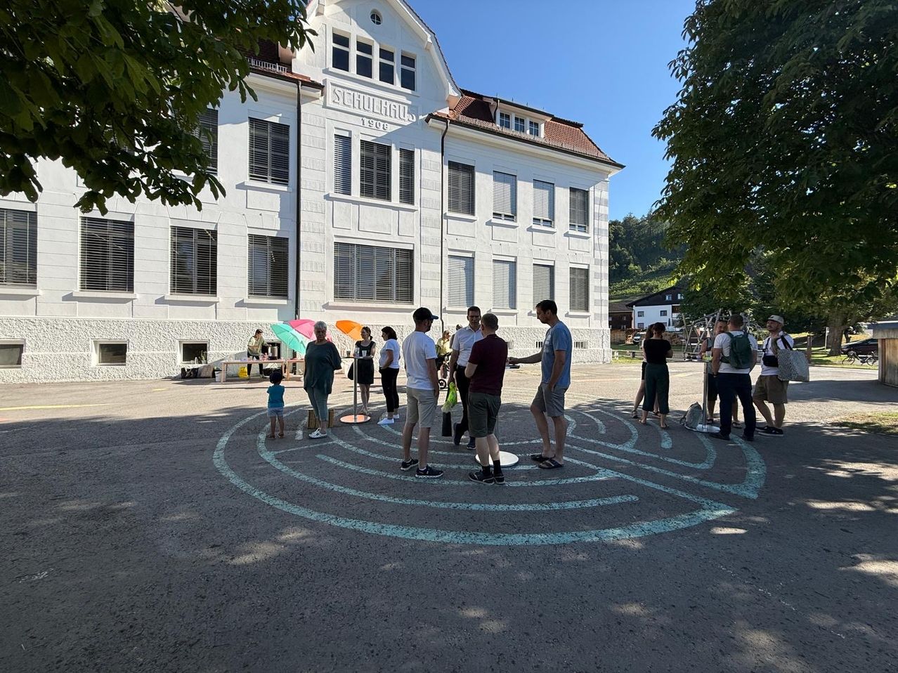A group of people stand in a circle in front of a white building with the name Schulhaus 1906 on it. The circle is painted on the ground.