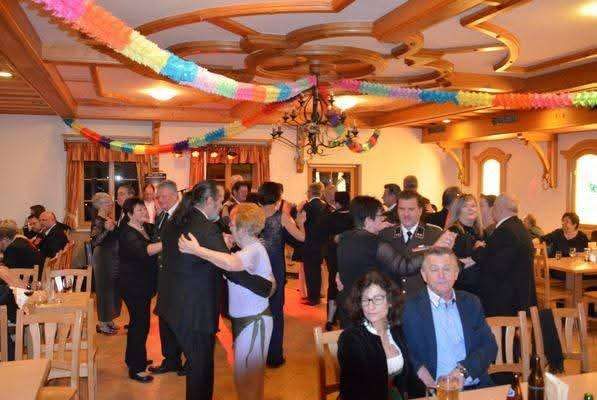 Eine große Gruppe von Menschen tanzt in einem Raum mit hölzerner Decke und Wänden. Es gibt Tische und Stühle, und eine Hängelampe hängt von der Decke.