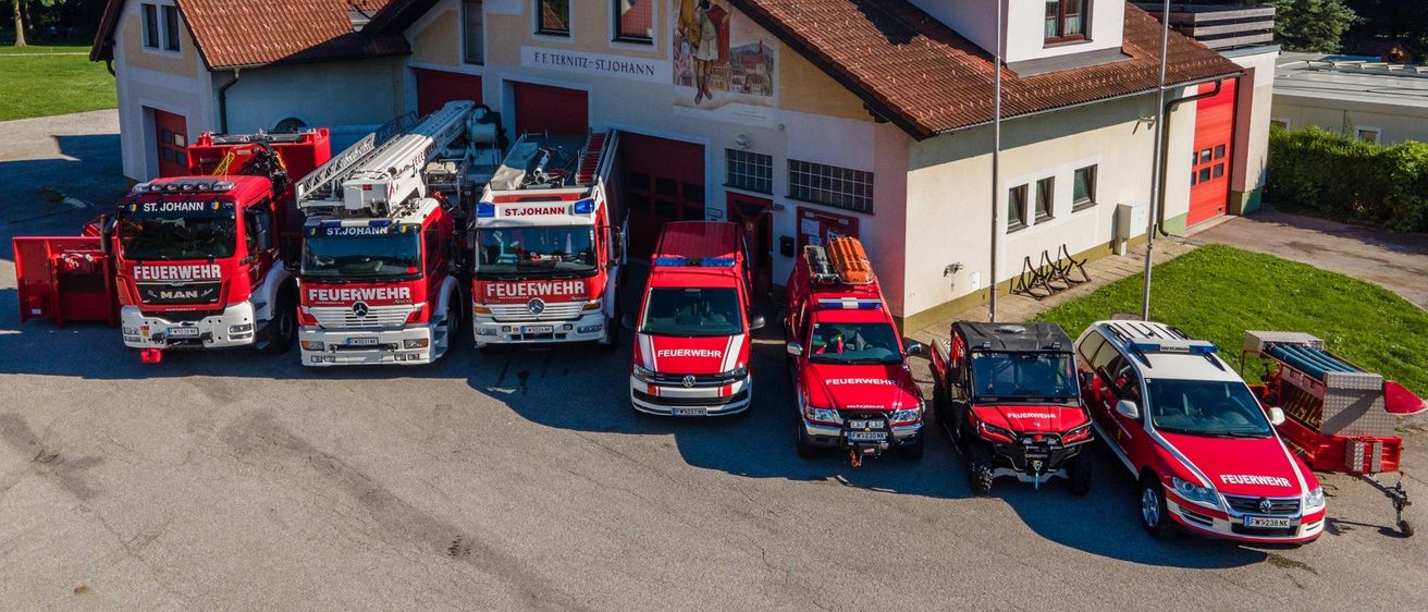 Ein Feuerwehrhaus mit rotem Dach und weißen Wänden ist zu sehen. Mehrere Feuerwehrfahrzeuge sind davor geparkt, alle mit 'Feuerwehr' gekennzeichnet. Das Gelände ist von Bäumen umgeben.