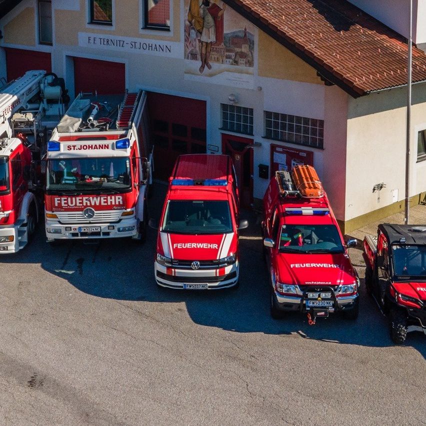 Ein Feuerwehrhaus mit rotem Dach und weißen Wänden ist zu sehen. Mehrere Feuerwehrfahrzeuge sind davor geparkt, alle mit 'Feuerwehr' gekennzeichnet. Das Gelände ist von Bäumen umgeben.