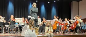Eine Gruppe von Kindern in Engelskostümen steht auf der Bühne und führt ein Musikkonzert auf. Geiger und Cellisten spielen hinter ihnen. Zwei Mädchen in Engelskostümen sitzen am Bühnenrand.