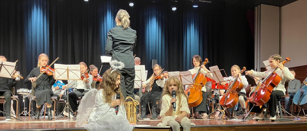 Eine Gruppe von Kindern in Engelskostümen steht auf der Bühne und führt ein Musikkonzert auf. Geiger und Cellisten spielen hinter ihnen. Zwei Mädchen in Engelskostümen sitzen am Bühnenrand.