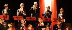 Eine Gruppe von Musikern spielt auf der Bühne mit Trompeten, Posaunen und anderen Instrumenten. Sie tragen formelle Kleidung und stehen vor einem dunklen Hintergrund.