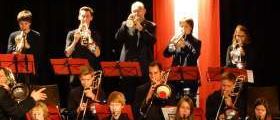 Eine Jazzband spielt auf der Bühne mit Trompeten, Posaunen und Saxophonen. Die Musiker sind in Schwarz und Rot gekleidet. Die Bühne ist mit roten Vorhängen und Notenständern ausgestattet.