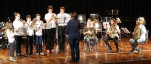 Eine Gruppe von Kindern und Jugendlichen spielt auf der Bühne, wobei sie Blech- und Holzblasinstrumente spielen. Die Kinder stehen, während die Jugendlichen sitzen. Der Dirigent steht vorne und dirigiert die Aufführung.