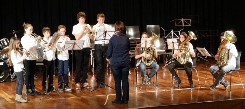 Eine Gruppe von Kindern und Jugendlichen spielt auf der Bühne, wobei sie Blech- und Holzblasinstrumente spielen. Die Kinder stehen, während die Jugendlichen sitzen. Der Dirigent steht vorne und dirigiert die Aufführung.