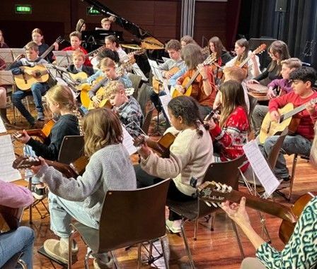 Eine Gruppe von Kindern und Erwachsenen versammelt sich in einem Raum für einen Musikunterricht. Sie spielen Gitarren, einige sitzen auf Stühlen. Es gibt Notenständer mit Musikblättern.