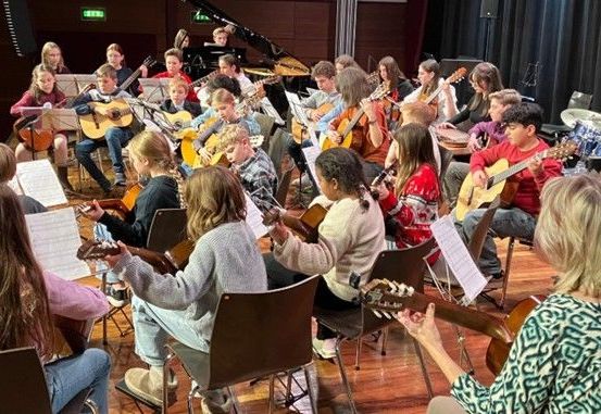 Eine Gruppe von Kindern und Erwachsenen versammelt sich in einem Raum für einen Musikunterricht. Sie spielen Gitarren, einige sitzen auf Stühlen. Es gibt Notenständer mit Musikblättern.