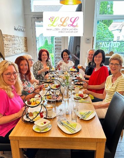 Eine Gruppe von Frauen sitzt an einem Tisch, lächelt und genießt ihr Essen, mit Tellern voller Speisen, Getränken und einem Schild mit 'LLL' im Hintergrund.
