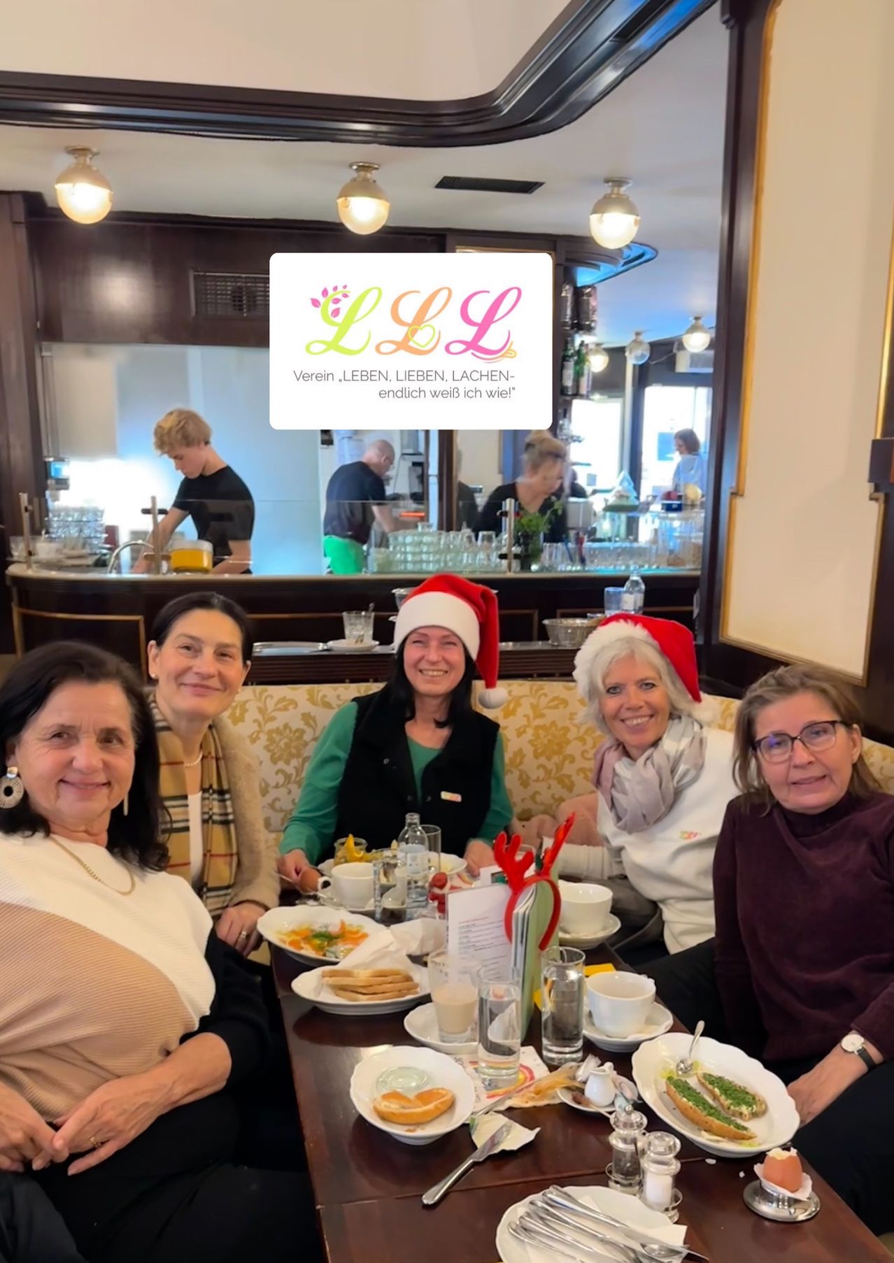 Fünf Frauen sitzen um einen Tisch in einem Restaurant. Sie tragen Weihnachtsmützen und lächeln. Hinter ihnen steht eine Person hinter der Theke. Im Hintergrund stehen mehrere Personen und ein Schild mit dem Text 'LLL' ist sichtbar.