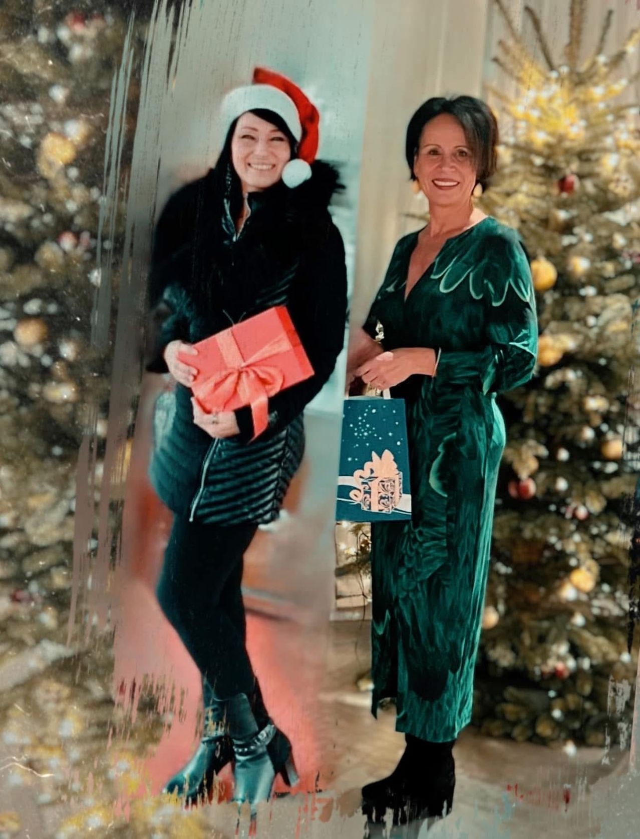 Zwei Frauen in Feiertagskleidung. Eine hält ein Geschenk, die andere einen Beutel. Sie stehen vor einem Weihnachtsbaum.