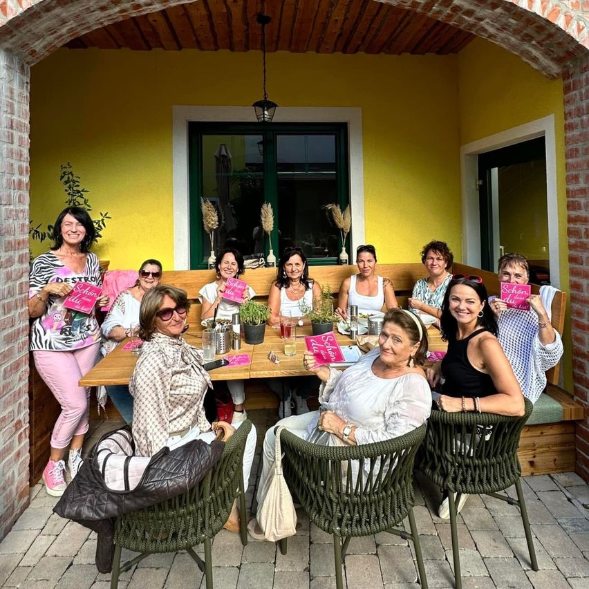 Eine Gruppe von Frauen sitzt um einen Tisch in einem gemütlichen Außencafe herum, alle halten rosa Bücher. Sie lächeln für ein Foto.