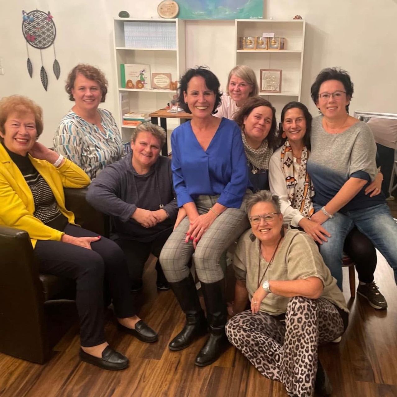 Eine Gruppe lächelnder Frauen in einem Wohnzimmer mit Holzboden, in verschiedenen Outfits gekleidet, posiert für ein Foto. Dahinter befinden sich Regale und eine Wand mit dekorativen Gegenständen.