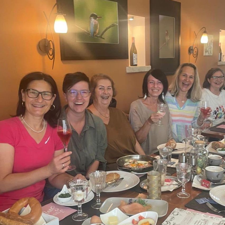 Fünf Frauen sitzen um einen Esstisch herum, lächeln und halten Weingläser. Der Tisch ist mit verschiedenen Gerichten gefüllt, darunter Eier, Brot und Obst. Eine Weinflasche und mehrere Lampen sind an der Wand hinter ihnen montiert.
