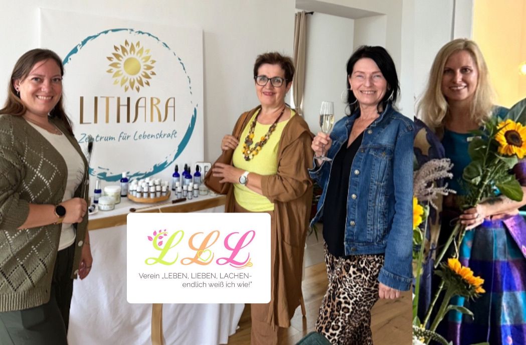 Vier Frauen stehen vor einem Tisch mit Produkten. Eine hält ein Glas Champagner. Ein Schild mit 'LITHARA' und einer Sonnengrafik ist hinter ihnen. Ein anderes Schild zeigt 'LLL' und 'Leben, Lieben, Lachen'.